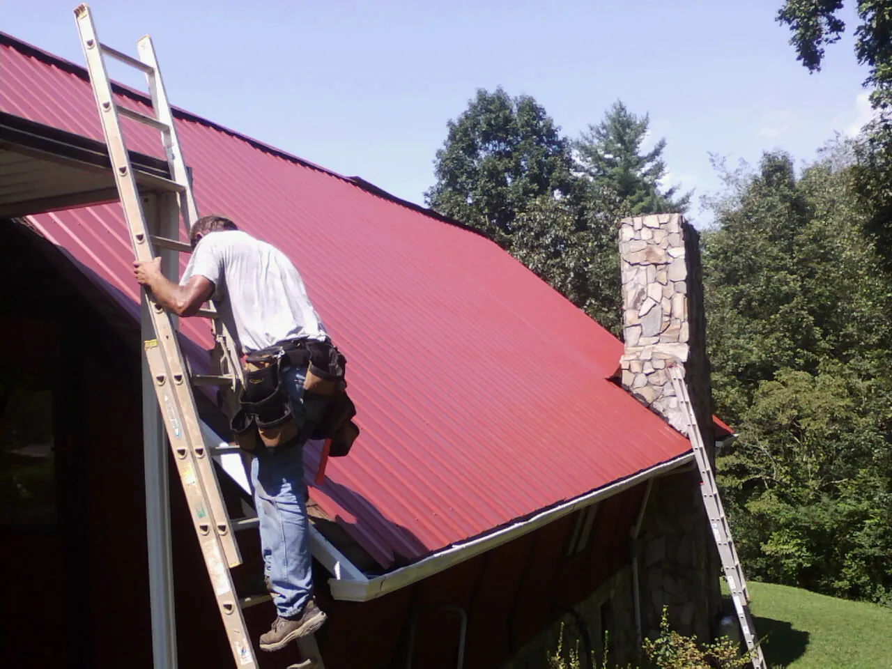 Roofer on a ladder beside a red metal roof and stone chimney — Soffit & Fascia Repair by Roofing Brothers in Canon